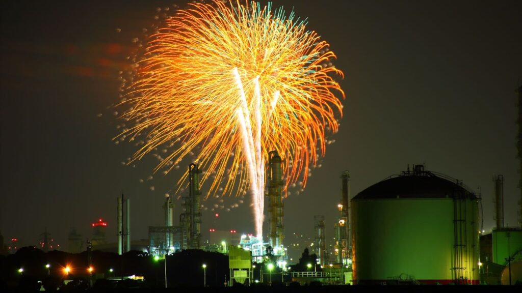 工業區建築與儲油槽上方綻放巨大的橘黃色煙火，與工廠的綠色燈光形成鮮明對比。