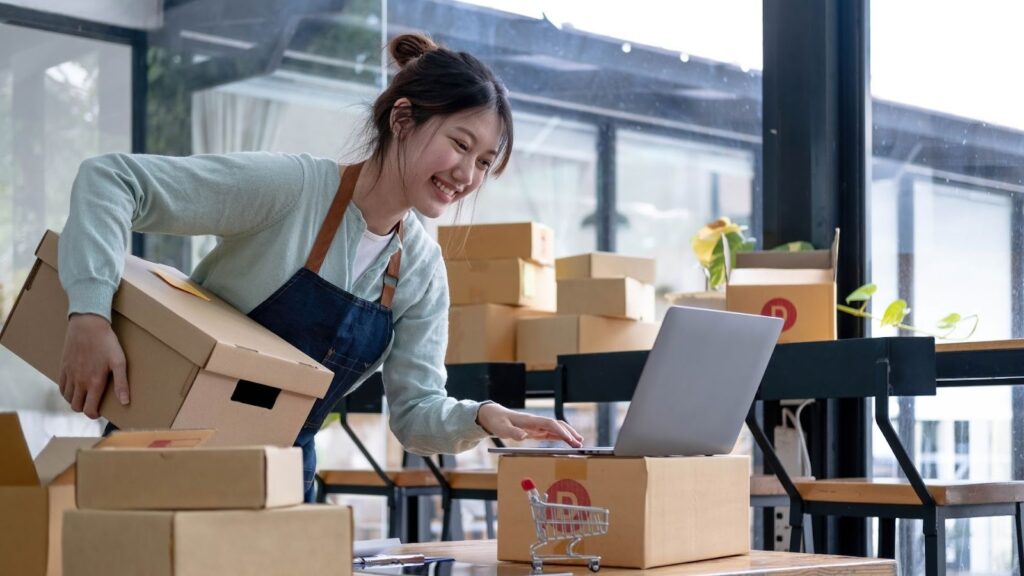 微笑的女創業者在工作室處理電商訂單，一手搬運紙箱並同時操作筆記型電腦，展現網拍經營、包貨與物流管理場景。