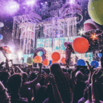 充滿活力的演唱會現場，眾多觀眾高舉雙手，舞台上佈滿燈光、彩帶、巨大的蘑菇道具和彩色大氣球