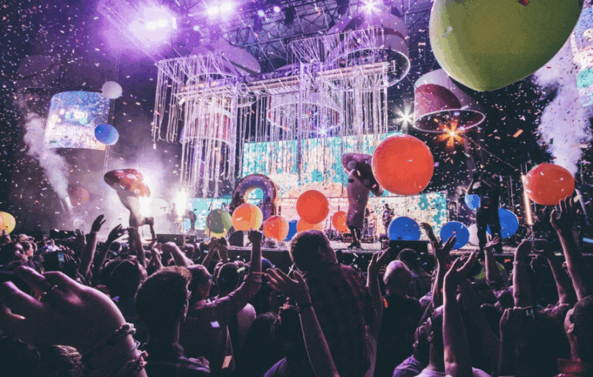 充滿活力的演唱會現場，眾多觀眾高舉雙手，舞台上佈滿燈光、彩帶、巨大的蘑菇道具和彩色大氣球