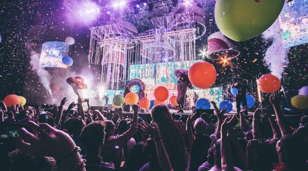 充滿活力的演唱會現場，眾多觀眾高舉雙手，舞台上佈滿燈光、彩帶、巨大的蘑菇道具和彩色大氣球