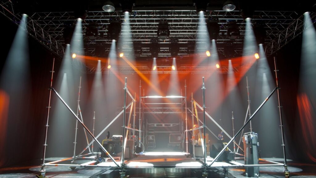 一個設有金屬桁架的舞台，從上方投射出多道白色的光束，中央交織著幾道橙色的燈光，營造出充滿霧氣的氛圍