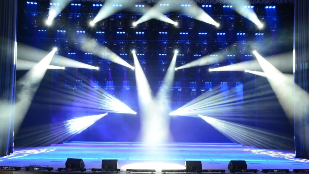 一個空曠的舞台，背後是藍色的幕布，無數白色的聚光燈束從上方投射下來，在地面上交織成複雜的光影圖案
