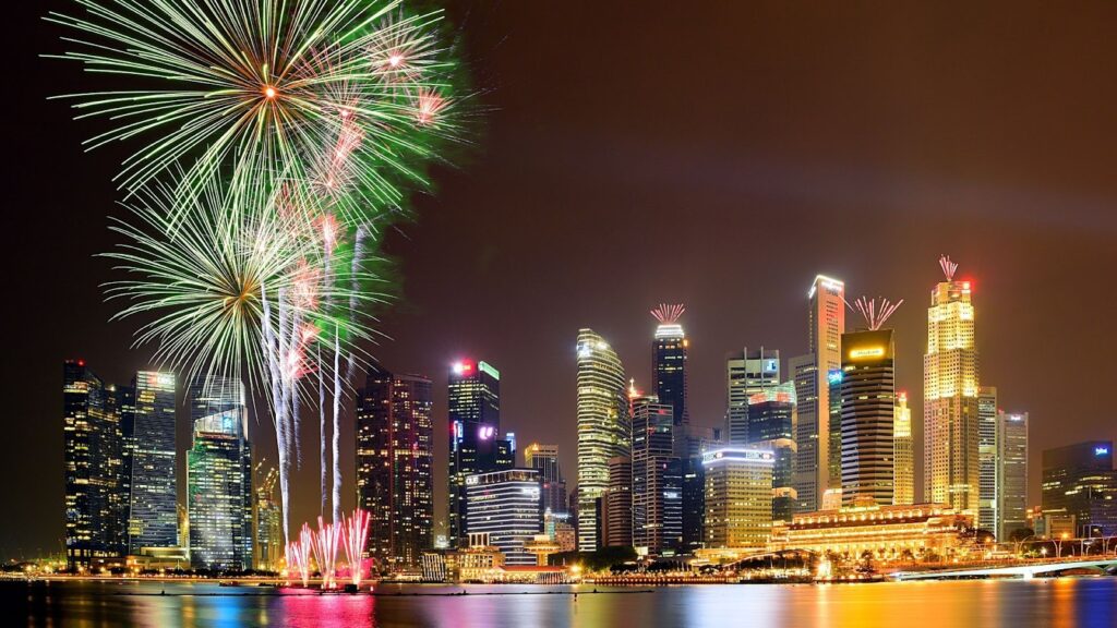 海灣城市夜景，右側是燈火通明的建築群，左側天空中綻放出巨大的綠色放射狀煙火，水面上閃耀著絢麗倒影