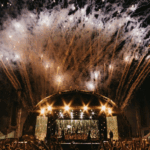 戶外音樂節表演現場，巨型拱形舞台散發金黃光芒，配合盛大的煙火特效與下方歡呼的群眾