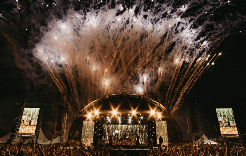 戶外音樂節表演現場，巨型拱形舞台散發金黃光芒，配合盛大的煙火特效與下方歡呼的群眾