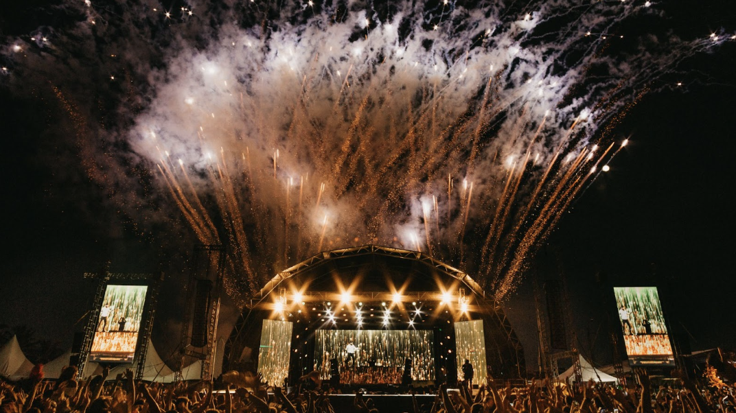 戶外音樂節表演現場，巨型拱形舞台散發金黃光芒，配合盛大的煙火特效與下方歡呼的群眾