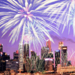 紫色與粉色調的夜空，城市現代建築上方有巨大的銀白色與青色煙火盛大綻放，並有細長的金色線條煙火交織