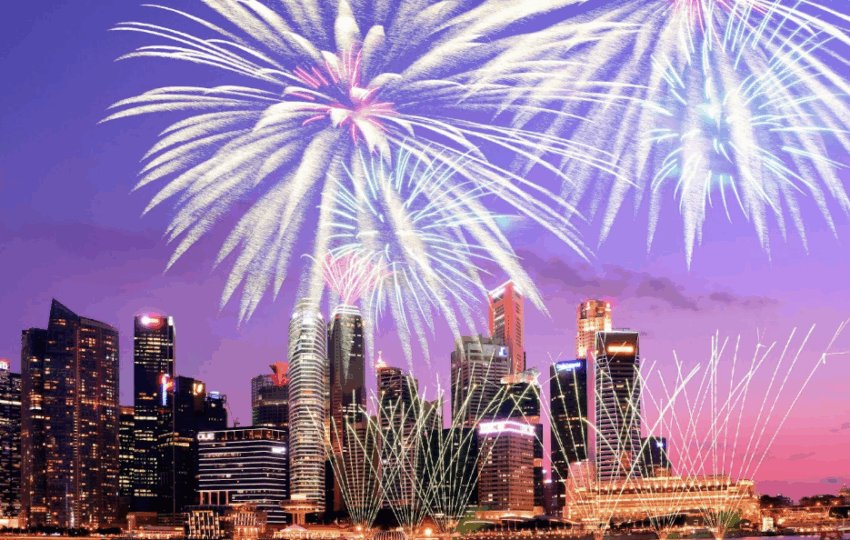 紫色與粉色調的夜空，城市現代建築上方有巨大的銀白色與青色煙火盛大綻放，並有細長的金色線條煙火交織