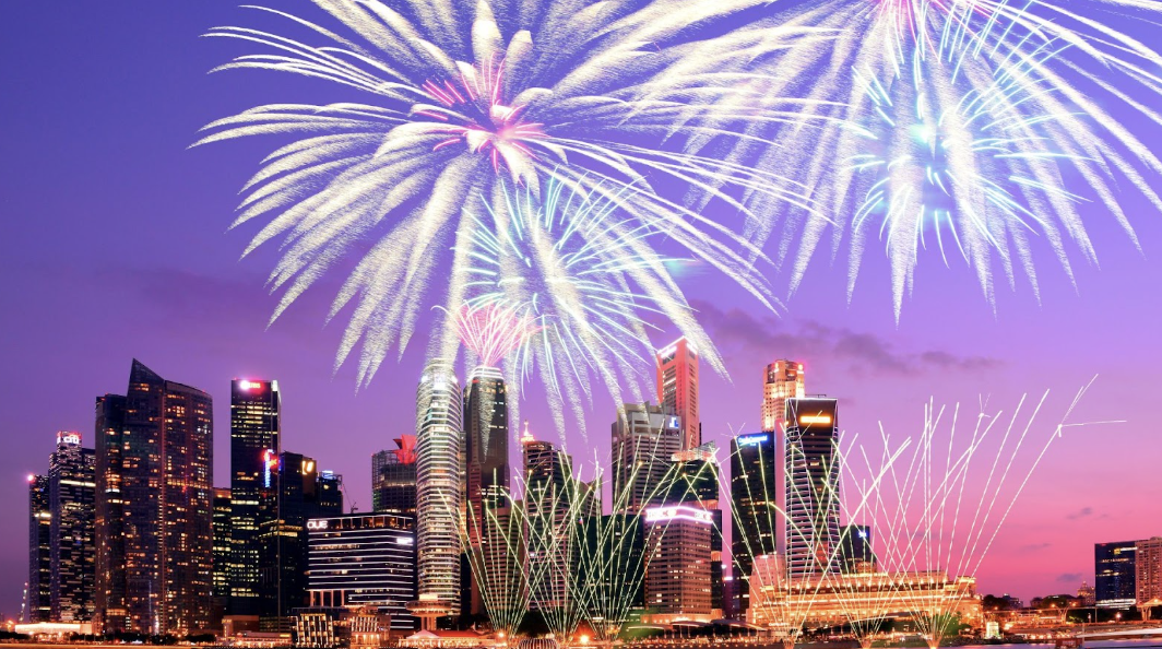 紫色與粉色調的夜空，城市現代建築上方有巨大的銀白色與青色煙火盛大綻放，並有細長的金色線條煙火交織